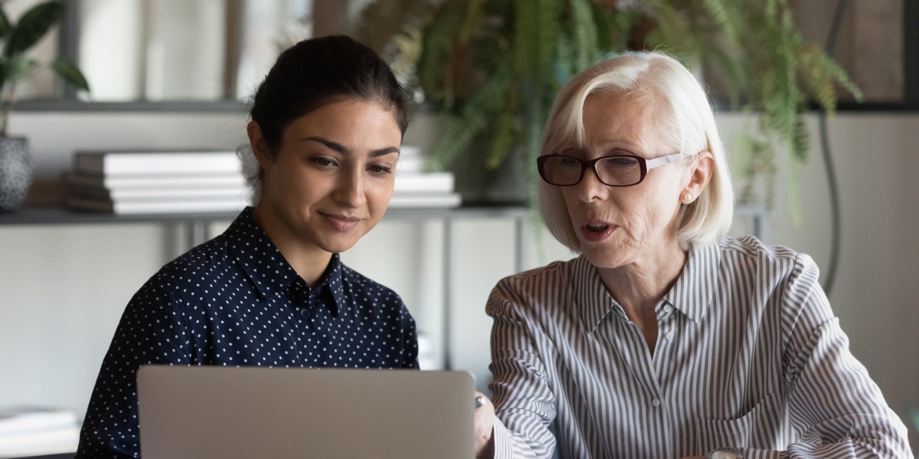 A young Asian woman with tied back dark hair and wearing a blue top, is sat looking at a laptop next to a white older woman with collar length white-bonde hair. She wears glasses and a light shirt. They are sat in an office environment in front of pl